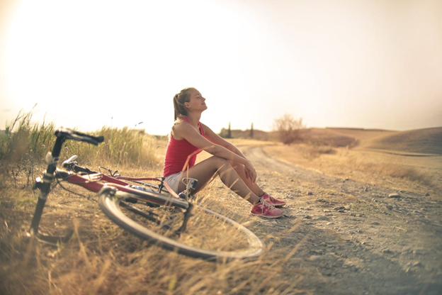 A woman in athletic clothes sits on a dirt path beside her bicycle, resting and enjoying the sunlight in a peaceful countryside setting.
